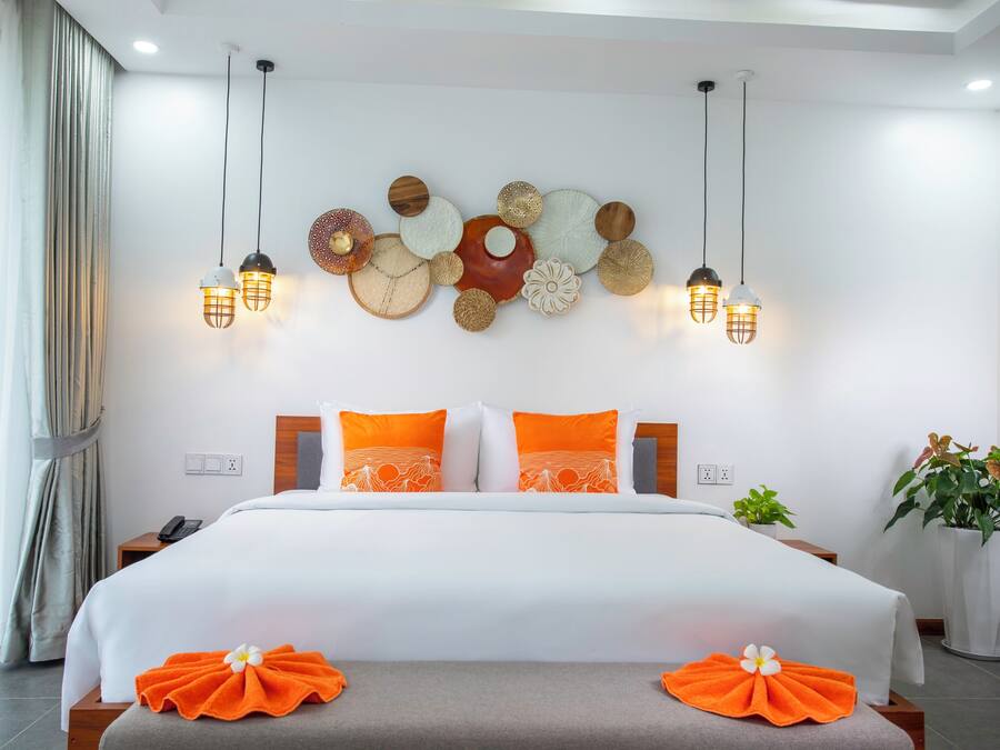 A hotel room with a large bed, orange pillows, a decorative wall piece, and pendant lighting.