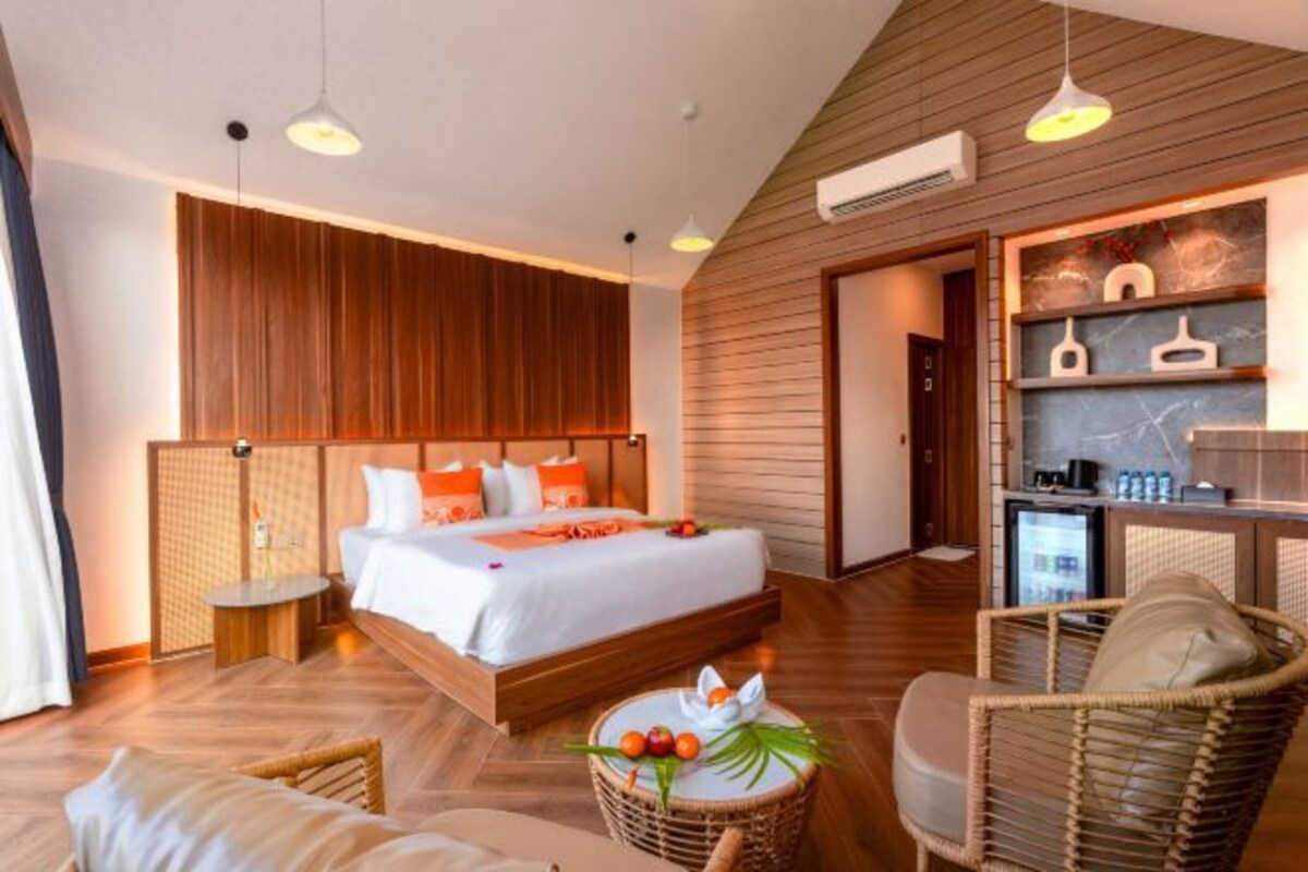 Quarto de hotel moderno com teto de madeira, cama grande com roupa de cama branca, pequeno kitchenette e móveis de vime.