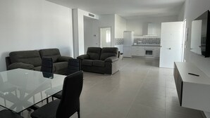 Uma sala de estar moderna com uma mesa de jantar de vidro, dois sofás e uma área de cozinha ao fundo.