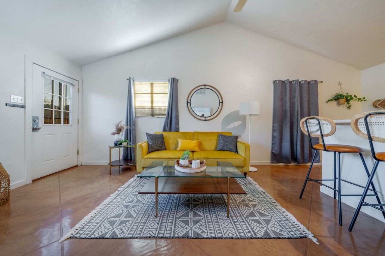 Un salon avec un canapé jaune, une table basse en verre, un tapis à motifs, un miroir rond et un coin repas avec des chaises en osier.