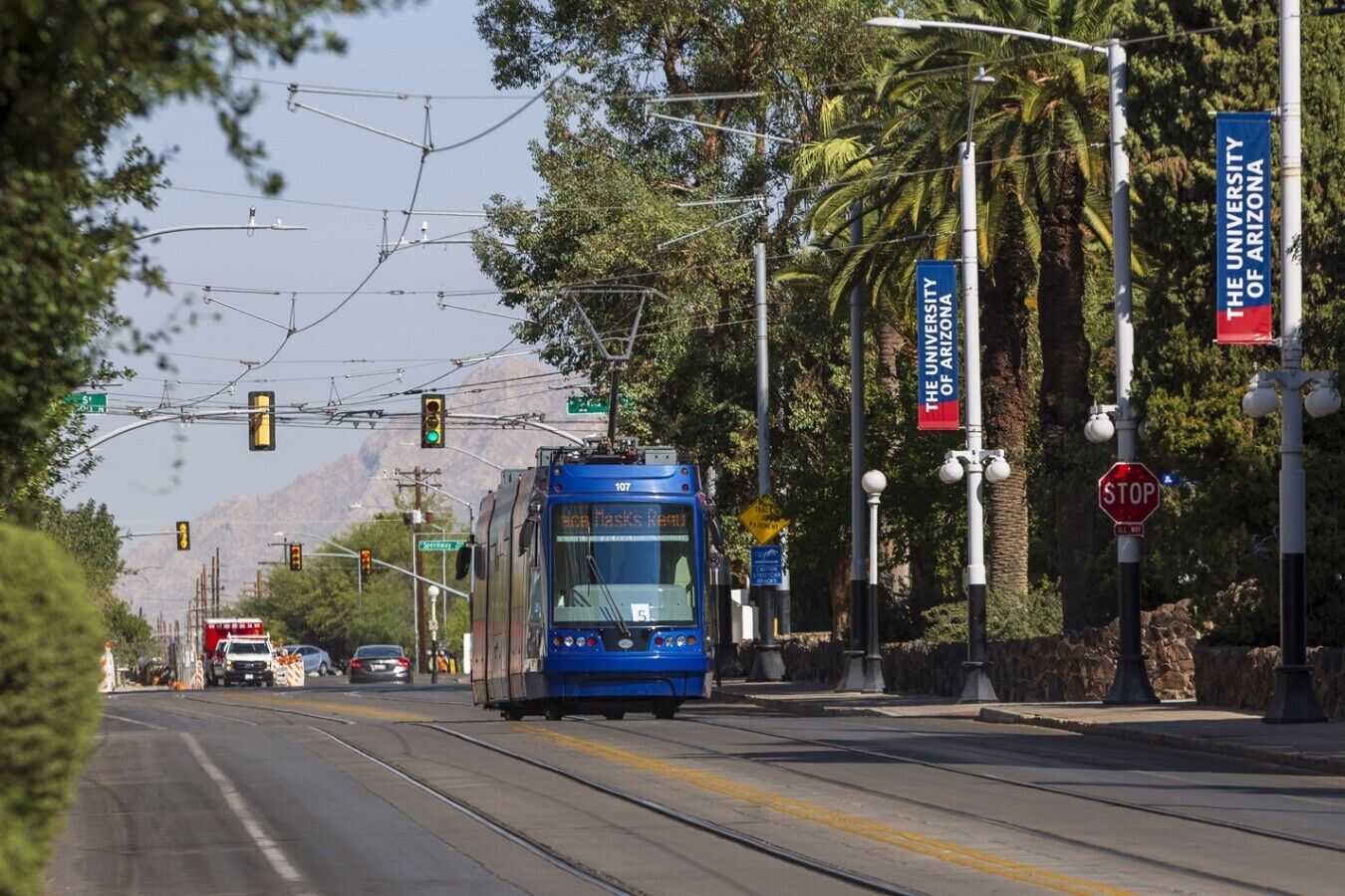A tram on a track with overhead wires, a stop sign, and a banner for the University of Arizona.