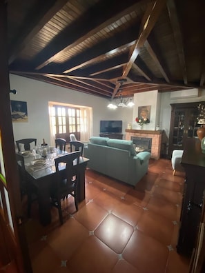 A dining area with a table set for four, a sofa, and a television in a room with a tiled floor and wooden ceiling beams. - Albarracín Rural House (Setenil de las Bodegas)