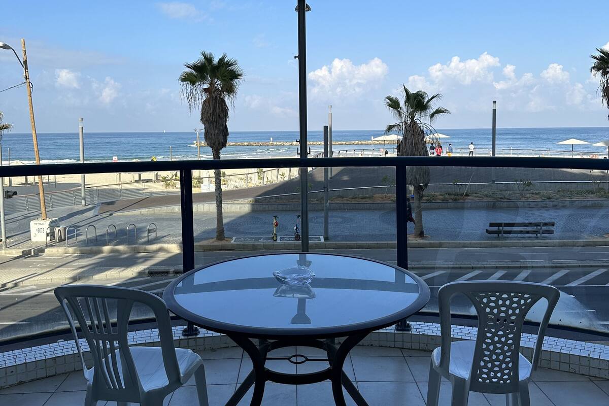 Une table ronde entourée de quatre chaises, placée sur une terrasse carrelée qui donne sur une plage bordée de palmiers et sous un ciel bleu azur.