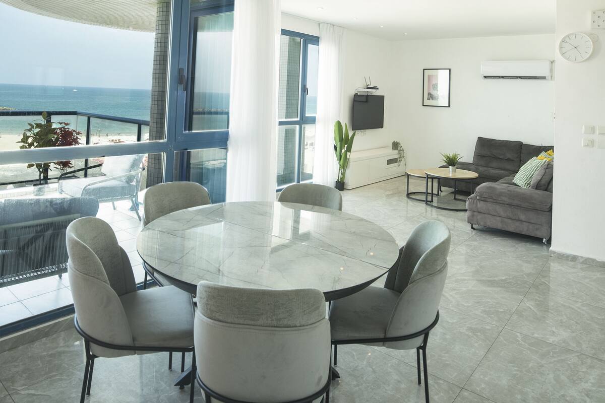 Un salon moderne avec une table ronde en verre et des chaises, un canapé gris et une vue sur la mer grâce à de grandes fenêtres.