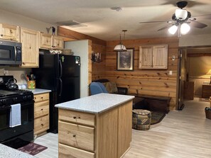 A compact kitchen with wooden cabinets, a black refrigerator, a microwave, and a stove. There is a ceiling fan and a small dining area with a table and chairs.