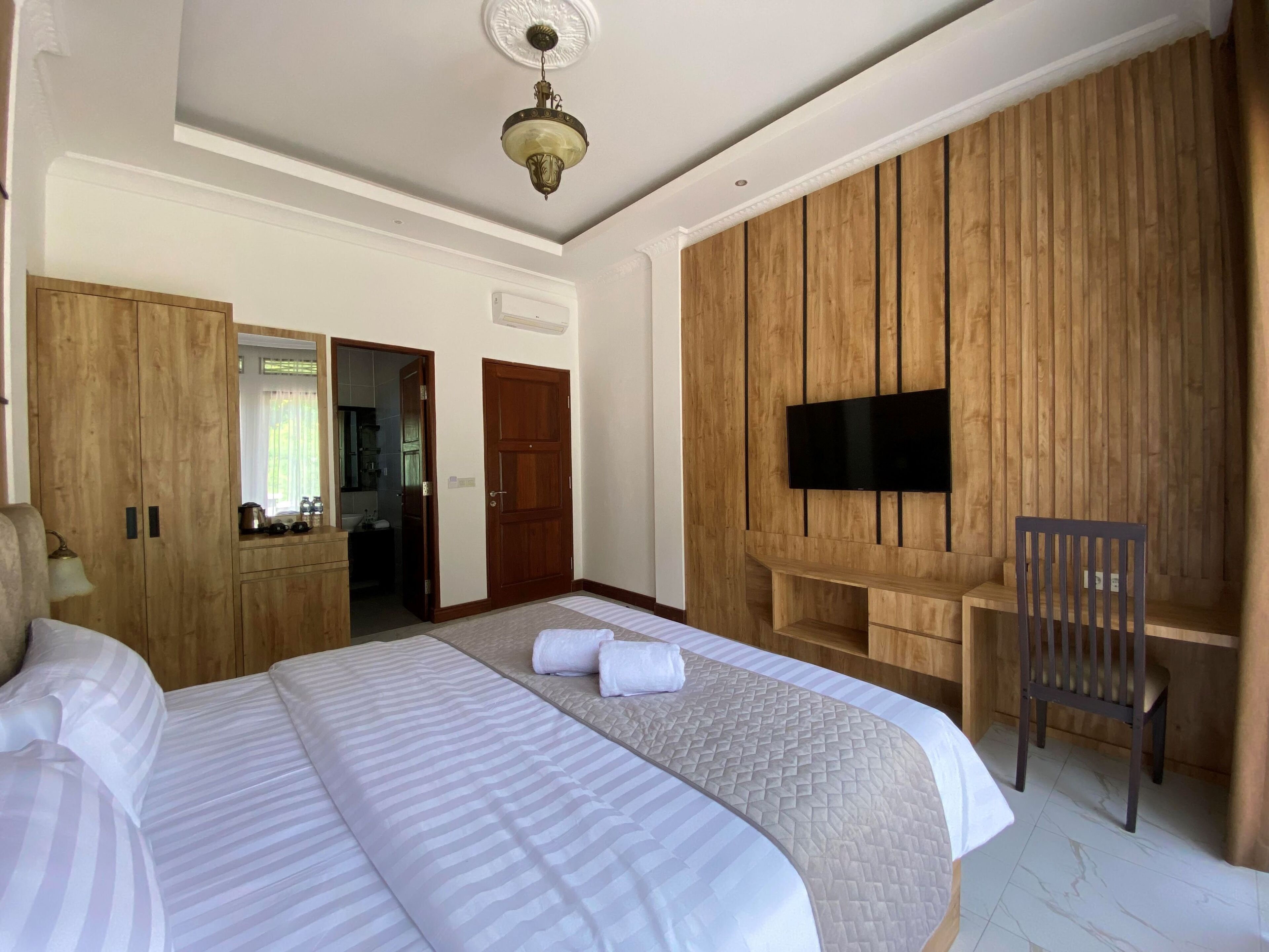 Quarto de hotel com uma cama grande, televisão fixada numa parede de madeira, uma cadeira e um guarda-roupa.