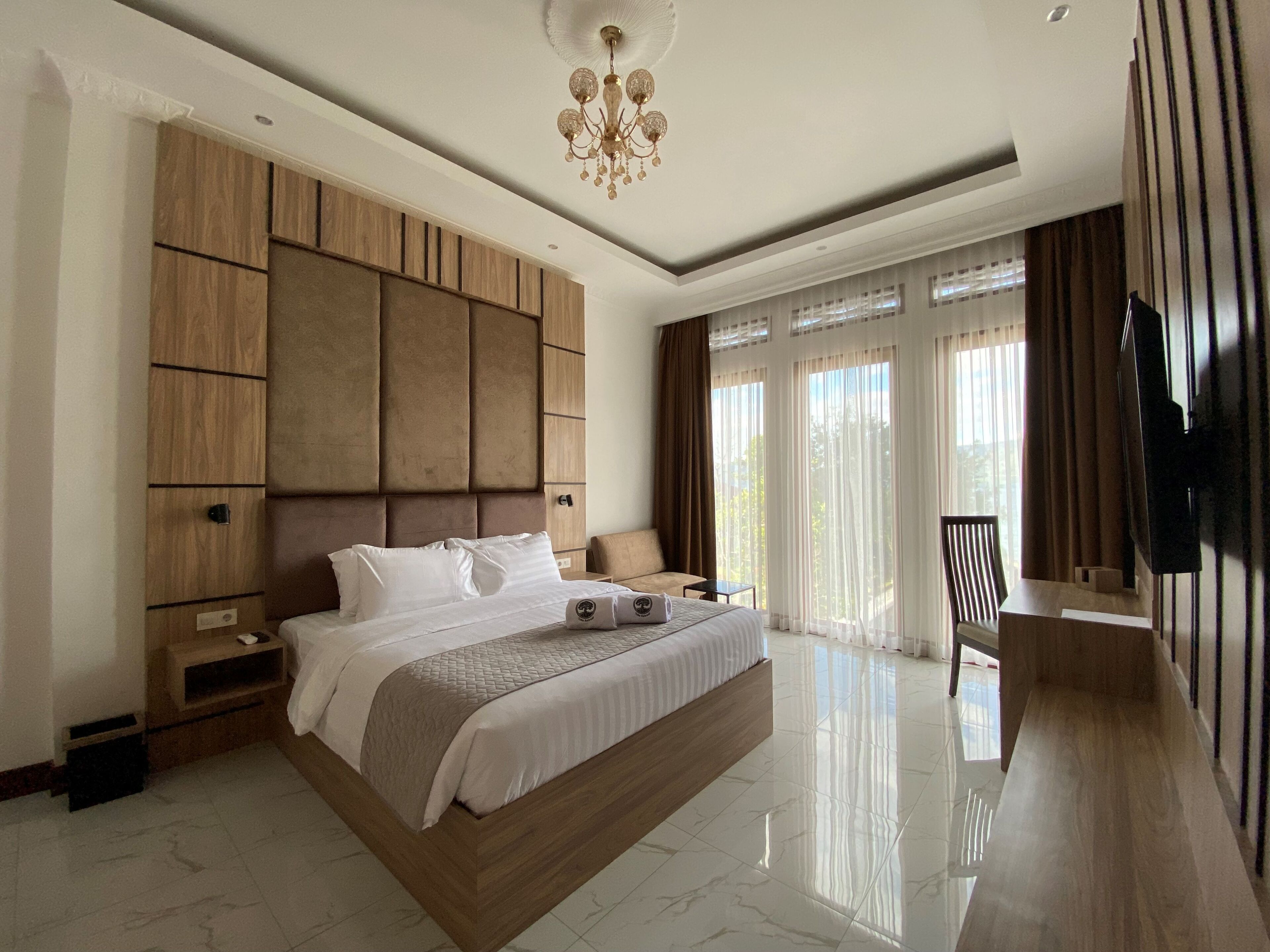 A modern bedroom with a large bed, a wooden headboard, a bedside table, a TV, and a balcony with curtains.