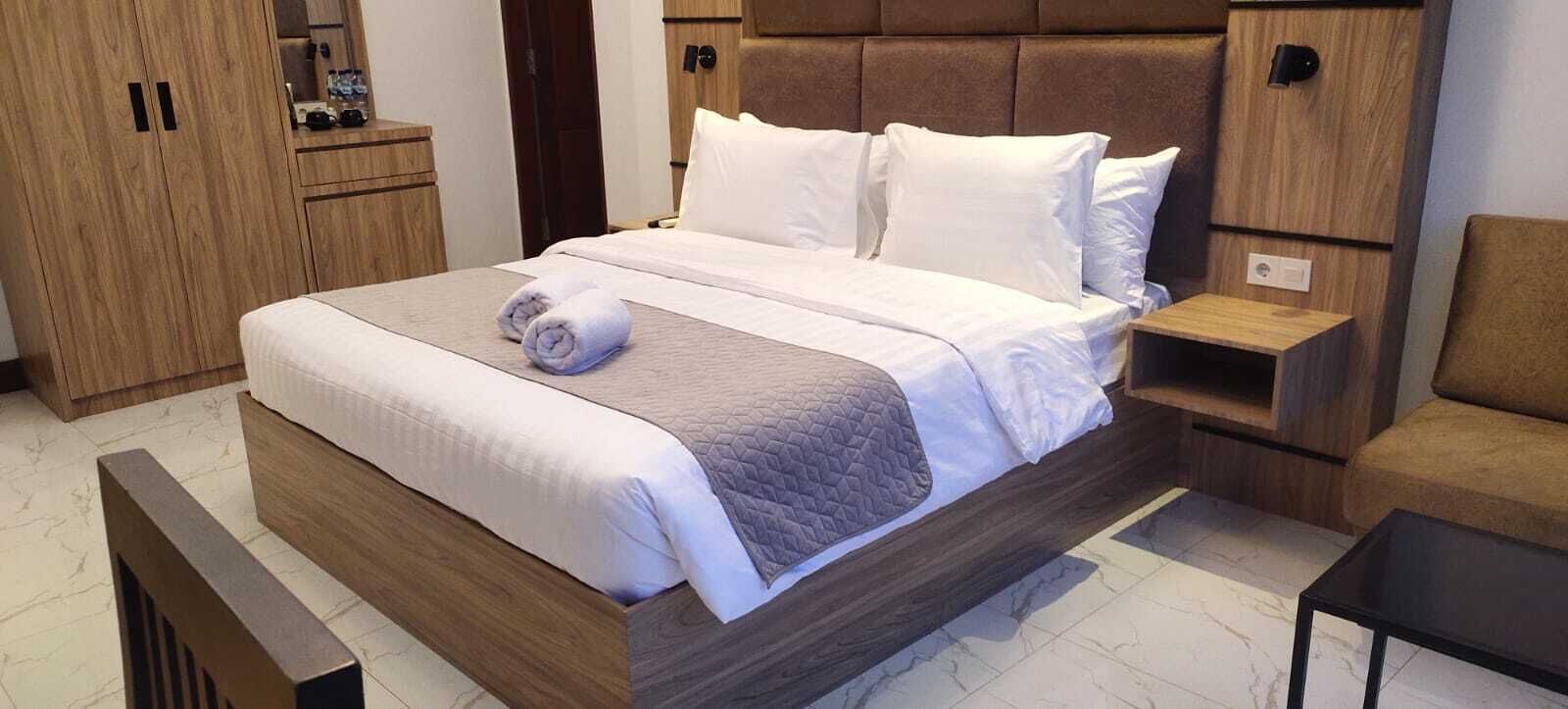 Ein ordentlich bezogenes Bett mit weißen Leinen und einem aufgerollten Handtuch, ein hölzerner Kopfteil, ein Holzschrank und ein kleiner Nachttisch mit einer Lampe.