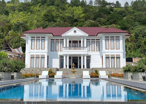 Un grand bâtiment blanc, surmonté d’un toit rouge, avec une piscine devant et une colline verdoyante en arrière-plan.