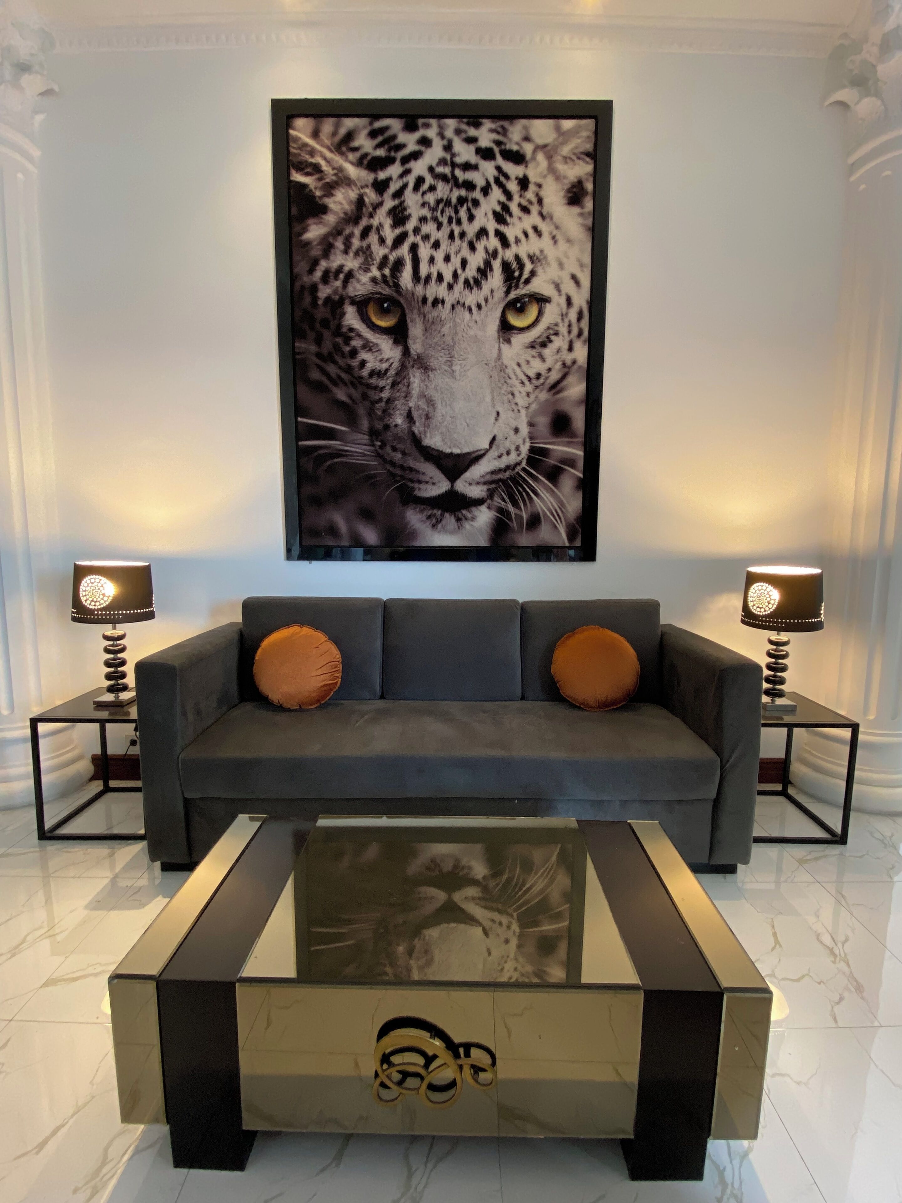 A modern living room with a grey sofa, two orange cushions, and a glass coffee table with a metal base.