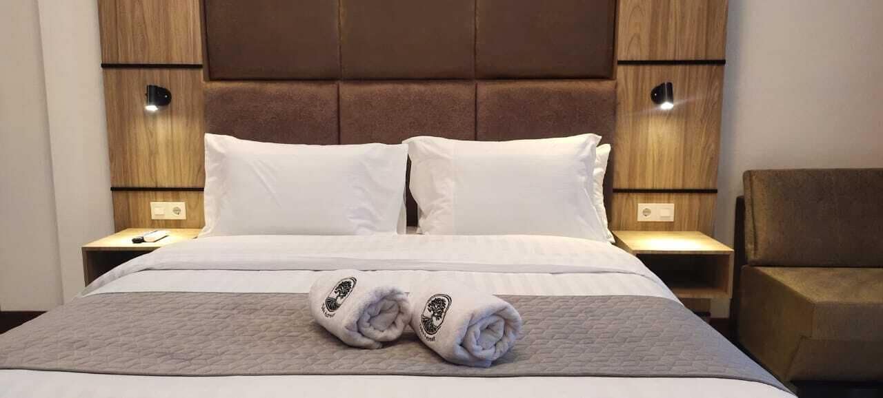 Habitación de hotel con una cama grande, dos almohadas blancas, un cabecero de madera y un par de toallas enrolladas sobre la cama.