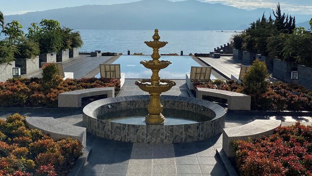 Ein Poolbereich mit einer zentralen Fontäne, Sitzmöglichkeiten und Blick auf das Meer und die Berge.