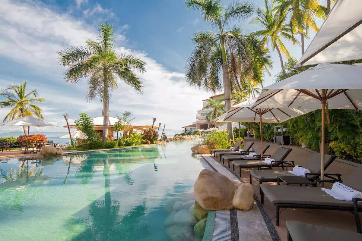 A pool area with lounge chairs and umbrellas, surrounded by palm trees and a clear sky.