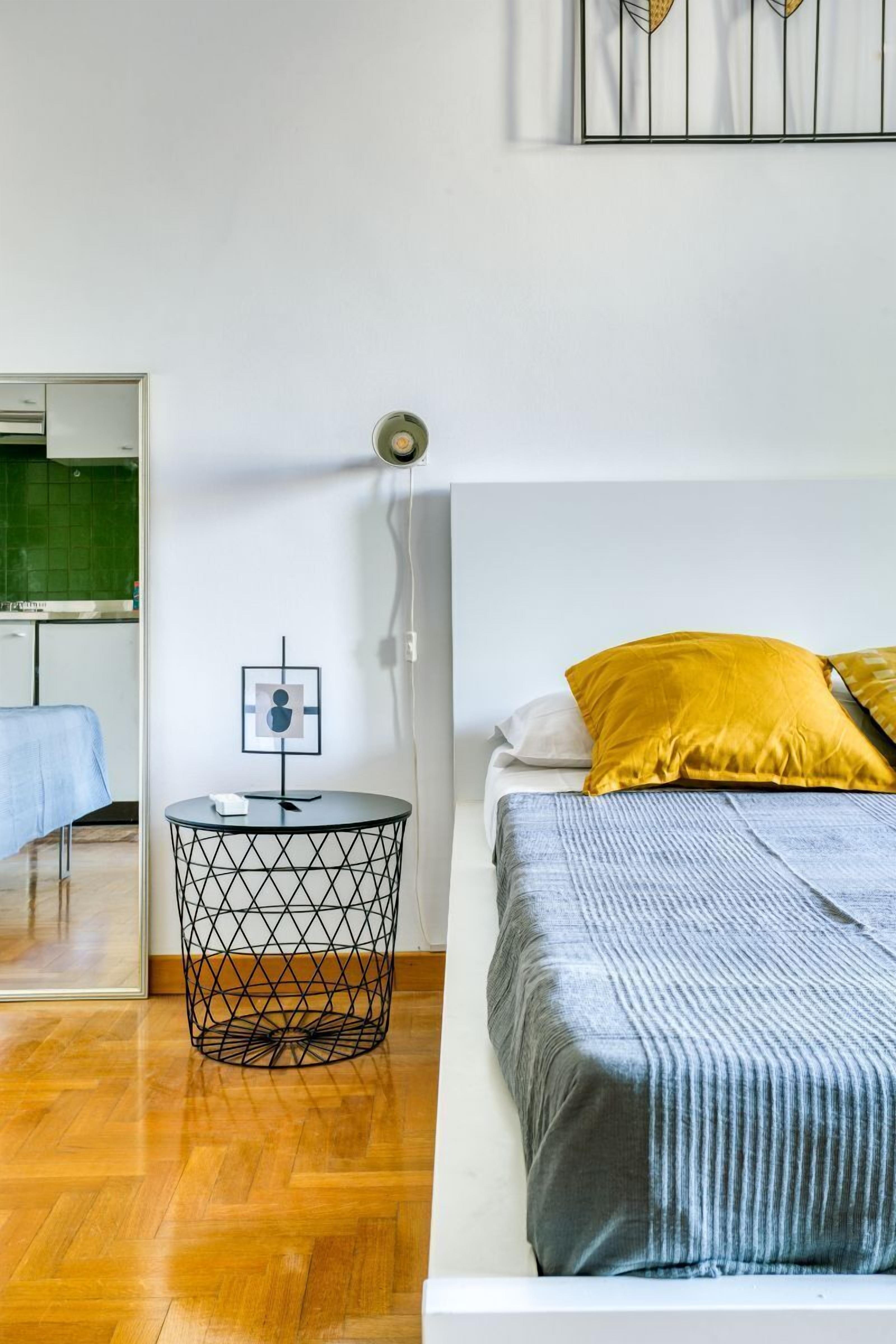 Quarto com uma cama, uma mesa redonda de arame e um luminário fixado na parede.