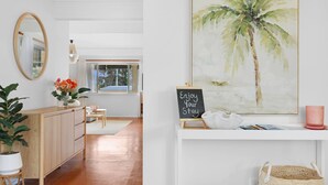 A modern room with a wooden cabinet, a mirror, a potted plant, a desk with a chalkboard sign, and a large painting of a palm tree.