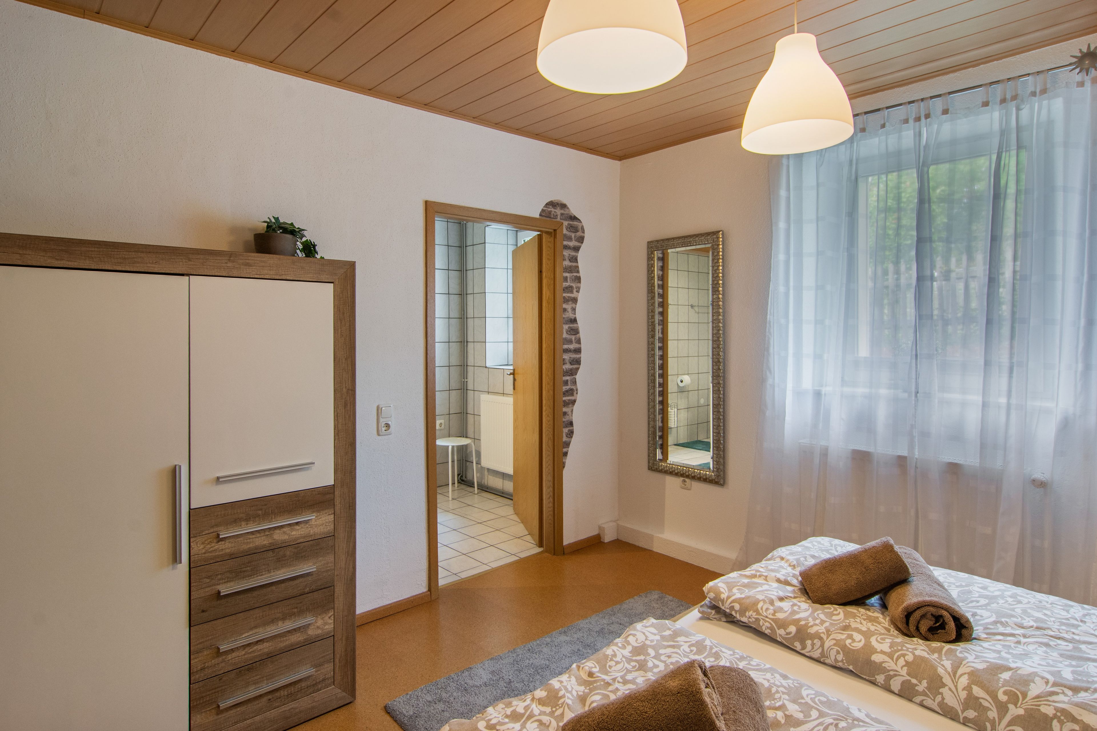 Une chambre avec une armoire, un miroir, un lit avec des serviettes et une fenêtre avec des rideaux légers.