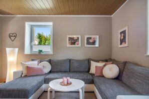 A modern living room with a sectional sofa, a small round table, and a window with a view of greenery. - Holiday Apartment "Fewo 2 - Osterberg" with Private Terrace, Shared Garden & Wi-Fi (Liebenau)