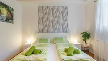 A hotel room with two beds, green striped bedding, a wall art piece, a potted plant, and a window with sheer curtains.