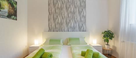 Habitación de hotel con dos camas, ropa de cama verde a rayas, una obra de arte en la pared, una planta en maceta y una ventana con cortinas de gasa.