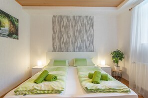 Quarto de hotel com duas camas, roupa de cama listrada em verde, uma obra de arte na parede, um vaso com planta e uma janela com cortinas translúcidas.