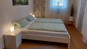 A bedroom with a bed, bedside tables, a nightstand, a window with curtains, a plant, and a painting on the wall. - Holiday Apartment "Fewo 2 - Osterberg" with Private Terrace, Shared Garden & Wi-Fi (Liebenau)