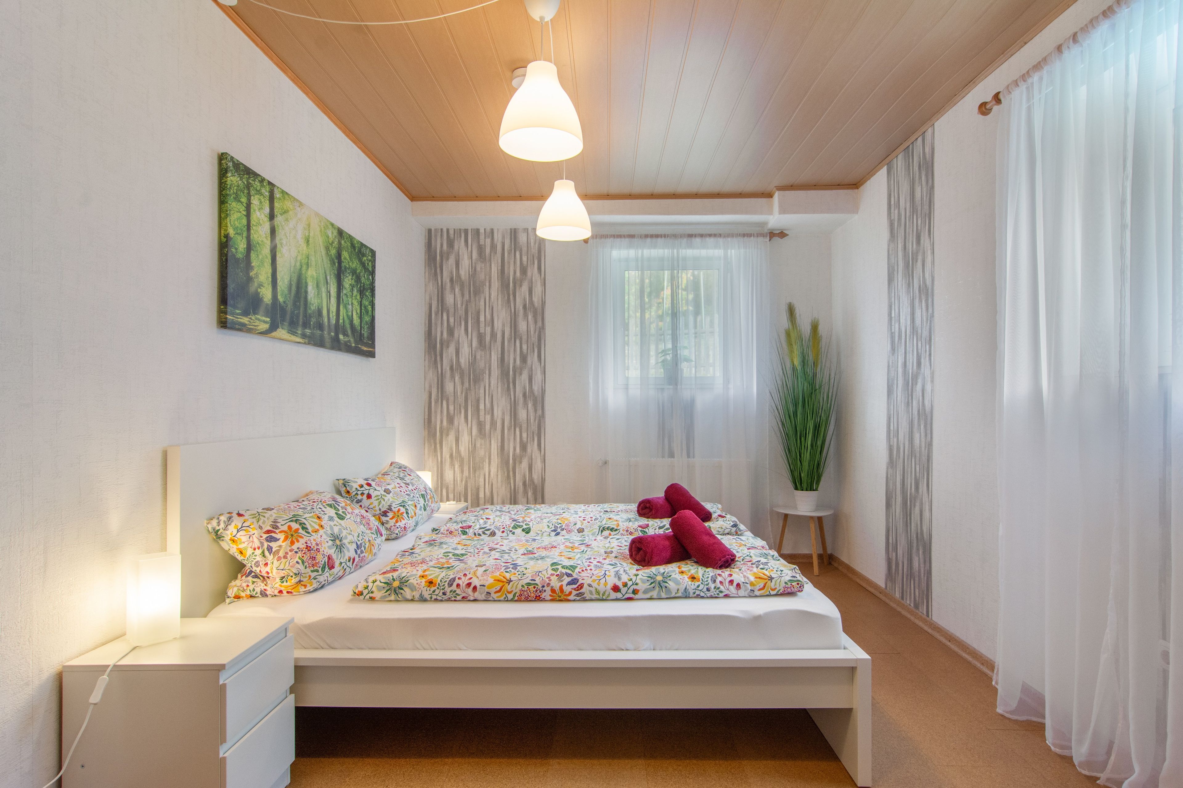 A bedroom with a bed, bedside tables, a nightstand, a painting, a window with curtains, and a potted plant.