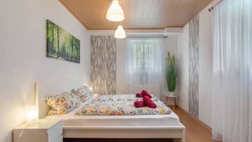 A bedroom with a bed, bedside tables, a nightstand, a painting, a window with curtains, and a potted plant.