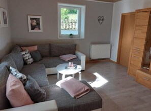 A living room with a grey sectional sofa, a small white table, a wooden wardrobe, and a window with a view of greenery.