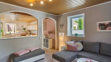 Ein modernes Wohnzimmer mit einem grauen Sofa, einem kleinen weißen Tisch und einem rosafarbenen Kissen. Durch einen Durchgang ist eine Küche zu sehen, und ein Fenster bietet Blick auf Grünflächen.