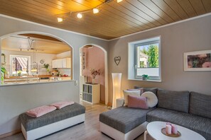 Un salon moderne avec un canapé gris, une petite table blanche et un coussin rose. On aperçoit une cuisine à travers une arche, et une fenêtre offre une vue sur la verdure.