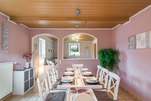 Un coin repas avec un plafond en bois, des murs roses et des meubles blancs. On aperçoit une plante sur la droite et un radiateur sur la gauche.