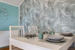 A dining table set with plates, bowls, glasses, and cutlery, against a wall with a tropical leaf pattern.