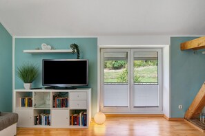 A living room with a flat-screen TV on a white stand, a bookshelf with books, a potted plant, and a large window with a view of greenery.