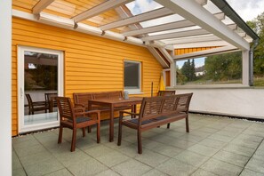 A covered patio with a wooden table and chairs, a glass door, and a view of the surrounding area.