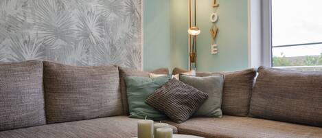 Un salon moderne avec un canapé d’angle, une table basse ronde ornée de bougies et un mur décoré avec le mot « AMOUR ».