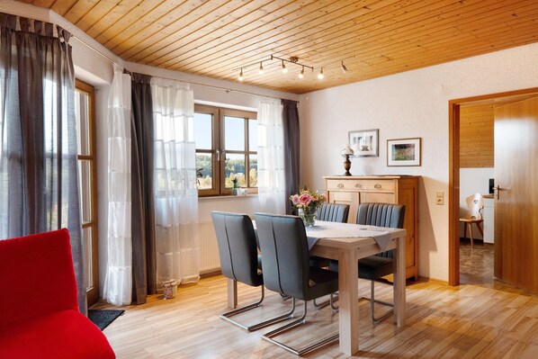 A dining area with a wooden table and chairs, a red chair, a wooden cabinet with a vase, and a view through a window. - Apartment 'Ferienwohnung Im Rosengarten' with Balcony and Wi-Fi (Ebenweiler)