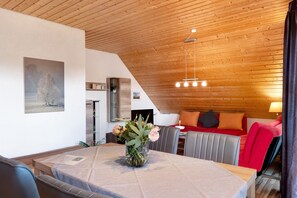 A modern interior with a dining table, chairs, a bed with pillows, a hanging light fixture, and a painting on the wall.