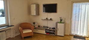 A room with a wicker chair, a TV mounted on the wall, a small cabinet, and a radiator.