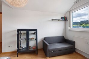 Ein Zimmer mit einem dunkelgrauen Sofa, einer Glastausch mit Spielzeug, einem weißen Heizkörper und einem Fenster mit Blick auf Grünfläche.