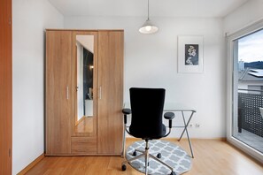 A room with a wooden wardrobe, a black office chair, a patterned rug, a ceiling light, and a window with a view of buildings.