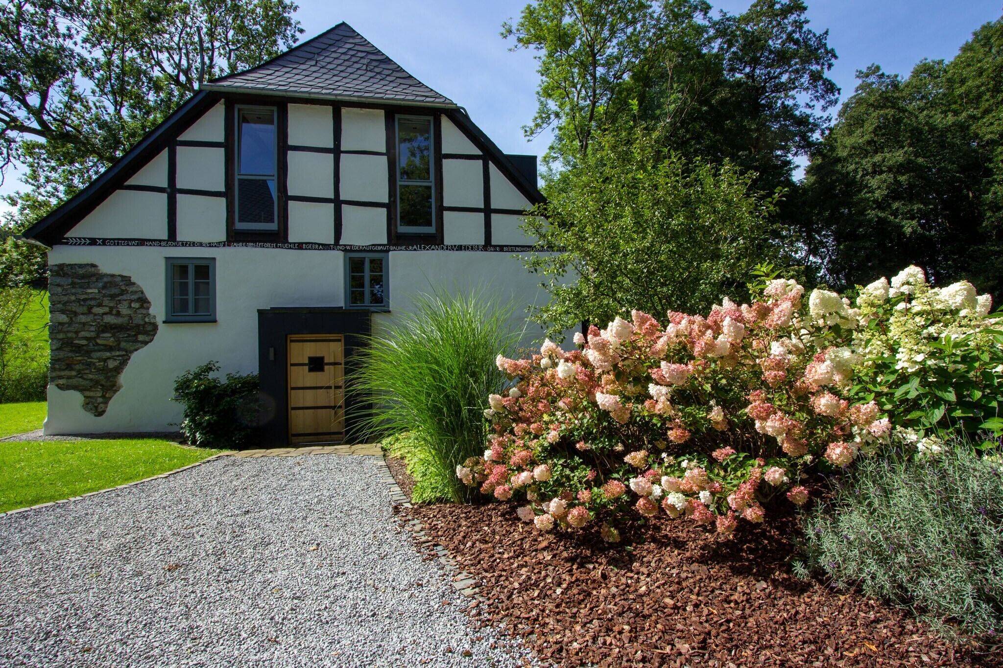 Uma casa em estilo Tudor com base de pedra, entrada por um caminho de cascalho e um jardim bem cuidado com flores cor-de-rosa e brancas.