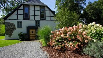 Uma casa em estilo Tudor com base de pedra, entrada por um caminho de cascalho e um jardim bem cuidado com flores cor-de-rosa e brancas.