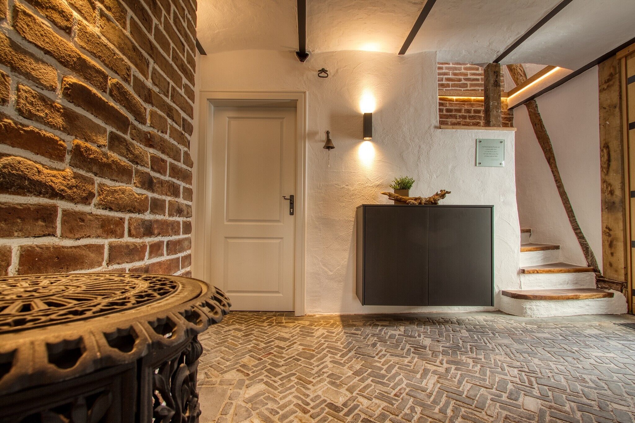 A hallway with a brick wall, a white door, a round table, a staircase, and a wall-mounted light fixture.