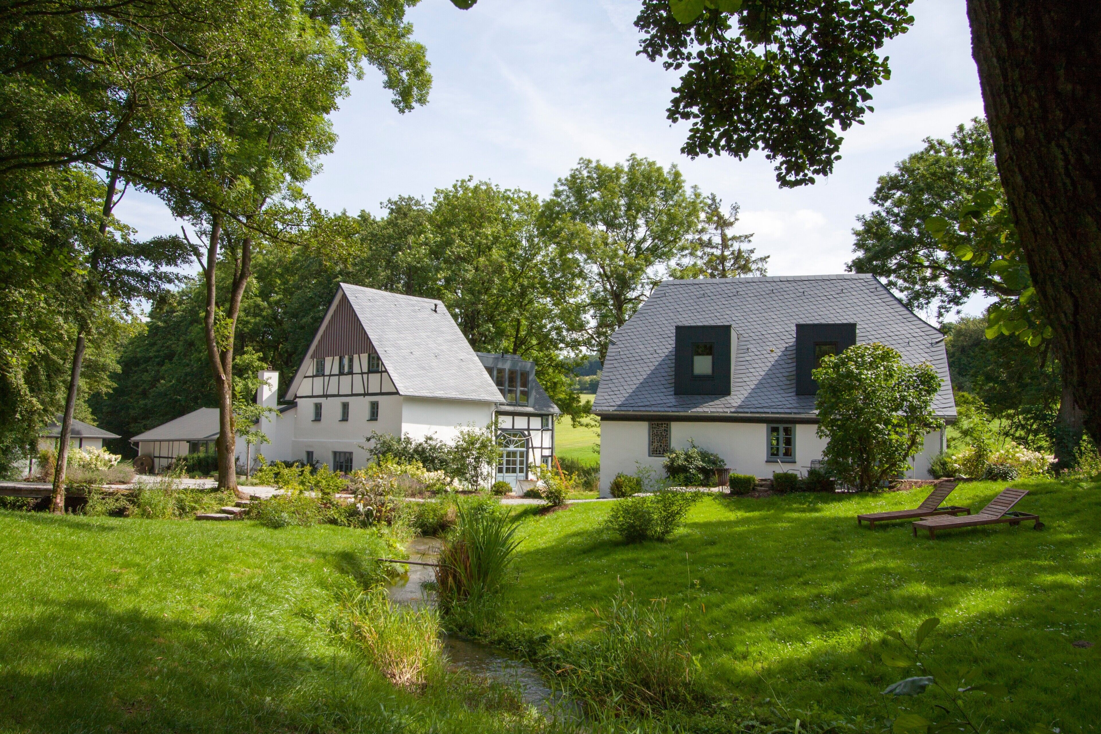Een traditioneel huis met een schuin dak, omringd door weelderige begroeiing en een kleine beek.