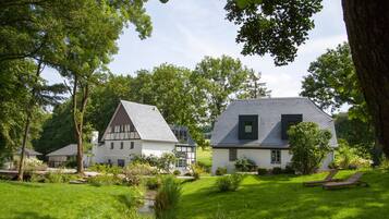 Een traditioneel huis met een schuin dak, omringd door weelderige begroeiing en een kleine beek.