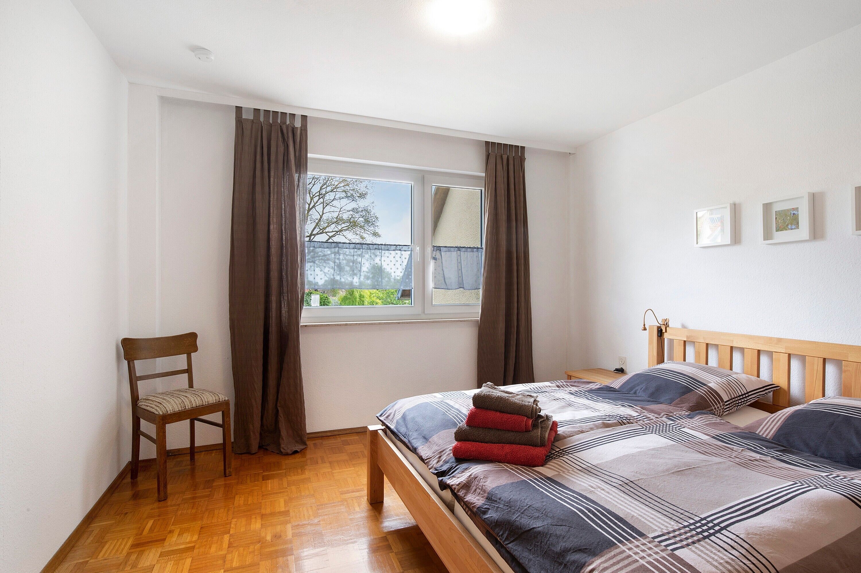 A bedroom with a wooden bed, a chair, a window with curtains, and a view of greenery outside.