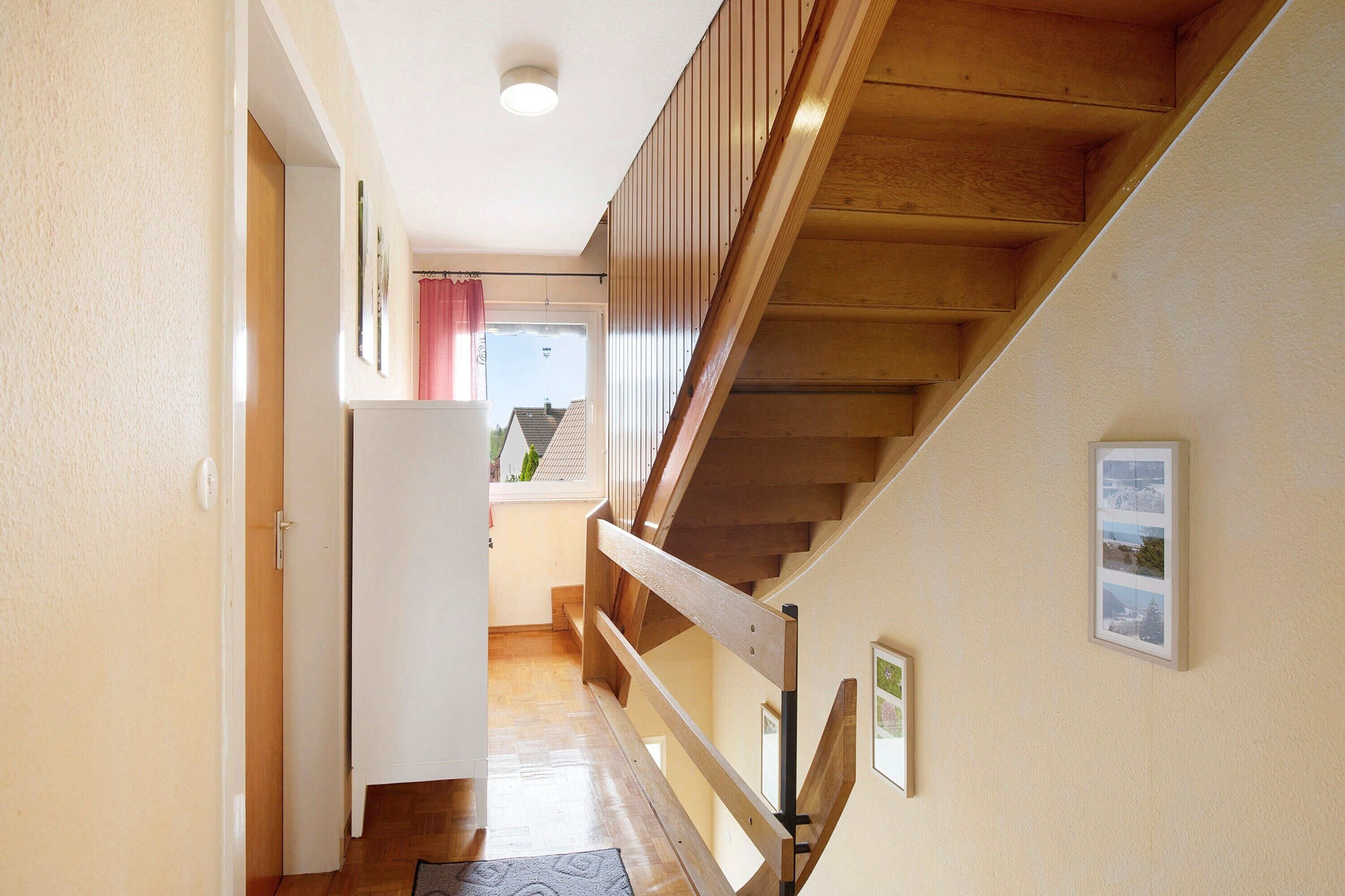 A wooden staircase with a handrail, a door on the left, and a window with a view of buildings outside.