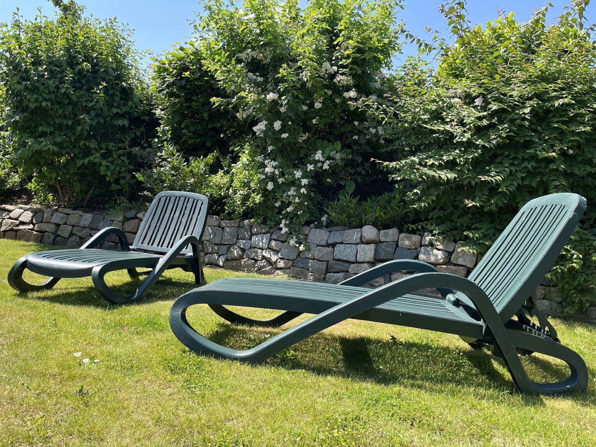 Two outdoor lounge chairs with a stone wall and lush greenery in the background.
