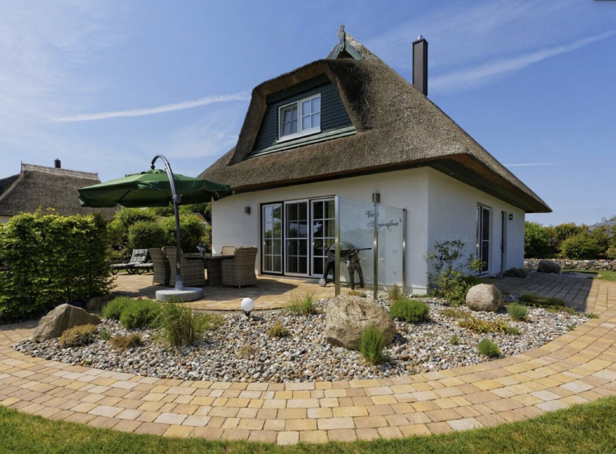 Une maison avec un toit de chaume, une terrasse avec des meubles d’extérieur et un jardin agrémenté de rochers et de plantes.