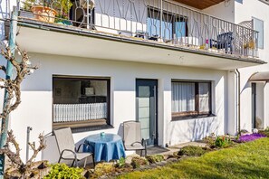 Un bâtiment à deux étages doté d’un balcon, d’un petit espace extérieur aménagé avec une table et des chaises, et d’un jardin fleuri.